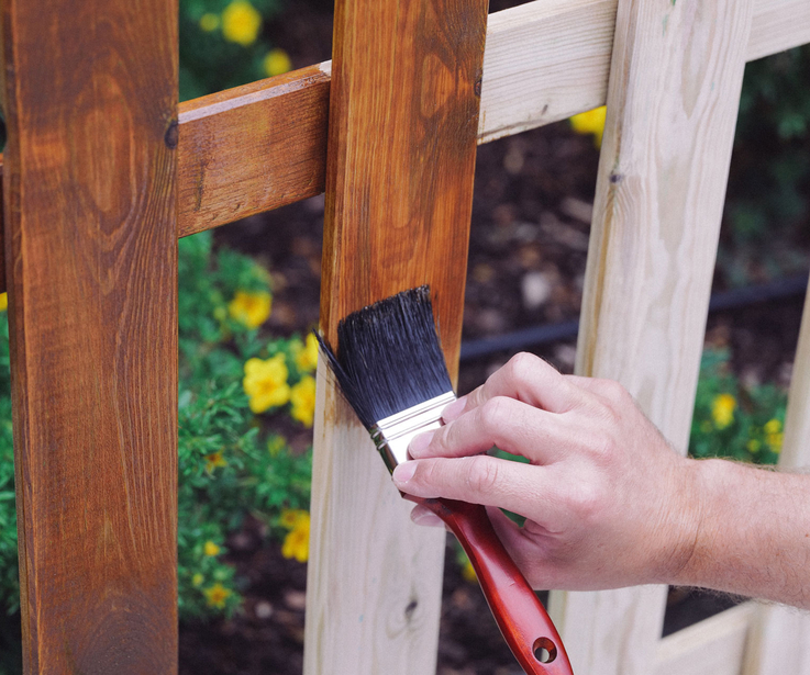 Ein Zaun wird mit einem Pinsel in brauner Holzlasur gestrichen