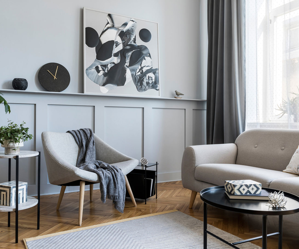 Blick in ein Wohnzimmer mit grau gestrichener Wand. Ein hellgraues Sofa steht vor einem hohen Fenster. Der Holzfußboden wirkt gemütlich. Schwarze Akzente wie eine Uhr, der Beistelltisch runden das Bild ab.