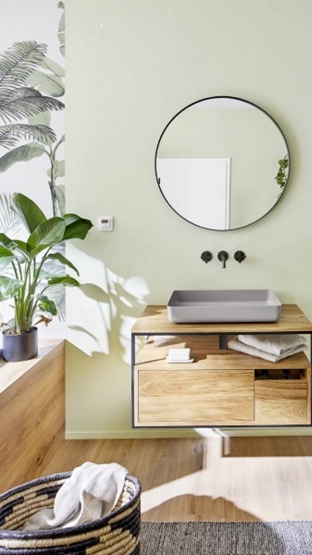 Hölzerner Waschtisch vor einer grünen Wand im Badezimmer. Ein runder Spiegel, ein Teppich und eine Pflanze runden die Gestaltung ab.