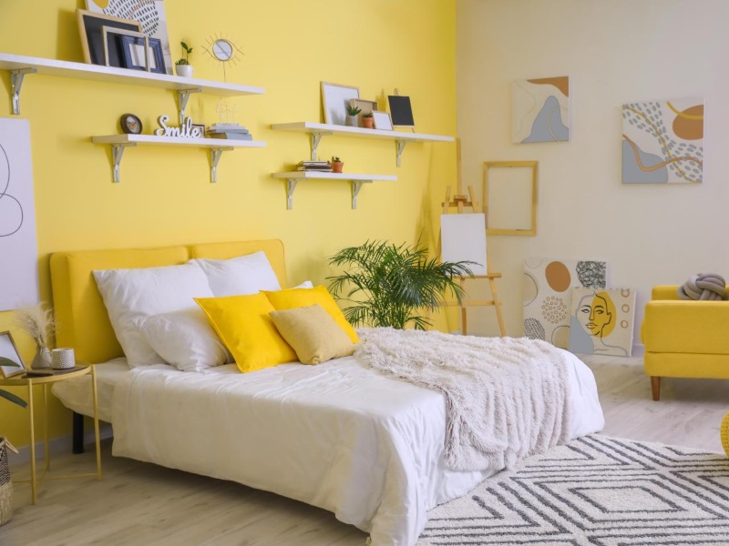 Ein Schlafzimmer mit gelber Wand, davor steht ein Bett. Das Zimmer ist gemütlich eingerichtet. Regale und Bilder an den Wänden und ein Teppich vor Bett wirken verspielt. Ein Sessel in Gelb und gelbe Kissen setzen tolle Akzente.